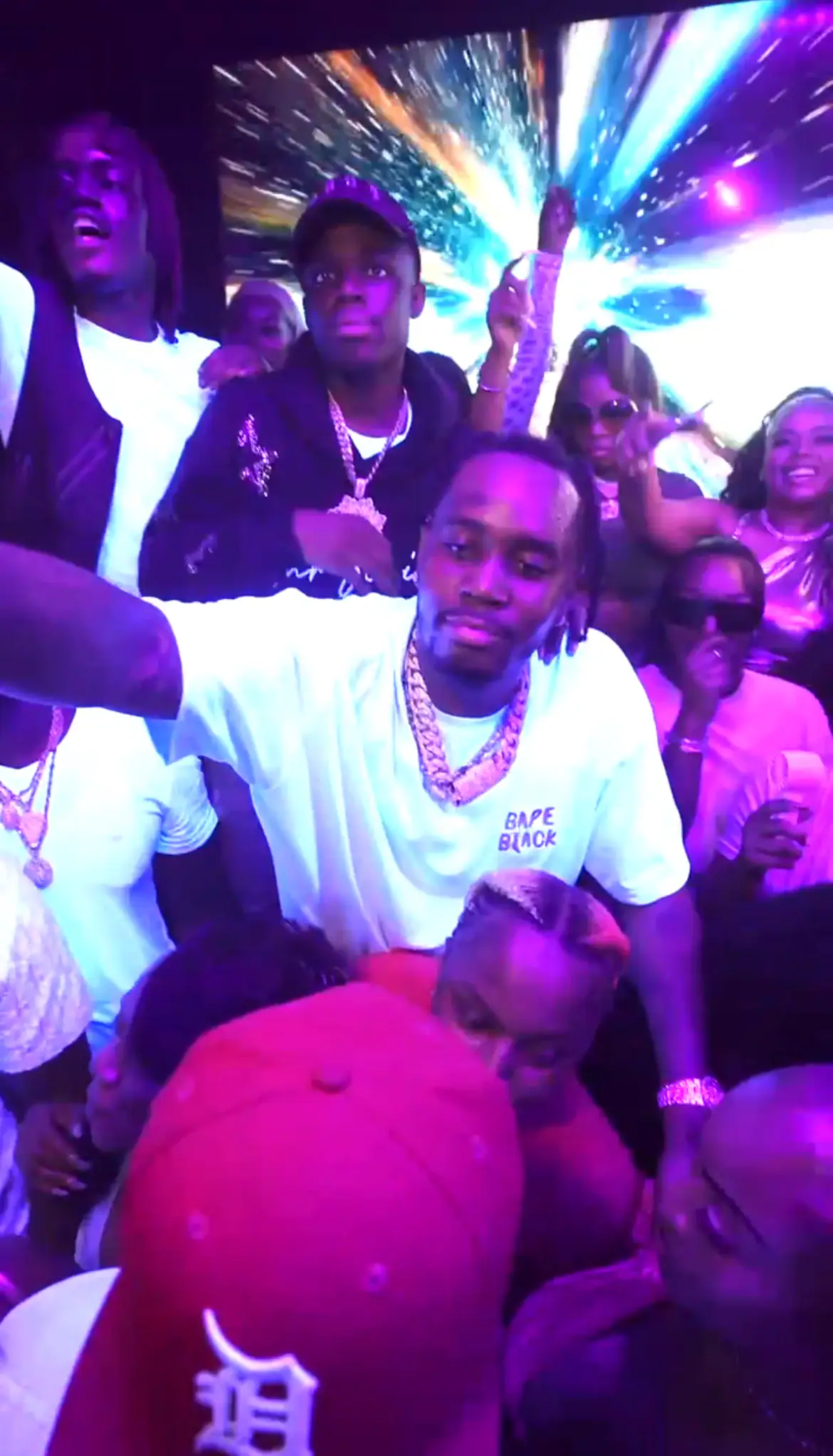 A group of people enjoy a lively party in a strip club with purple lighting. The central figure, wearing a white shirt and gold chains, poses amid cheering friends as a vibrant screen with bright colors glows in the background.