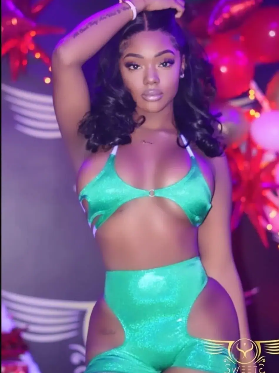 A woman poses confidently in a shiny green cutout outfit, with one hand raised to her head. The background, reminiscent of a vibrant strip club, features colorful balloons and blurred decorative elements in pink and red tones.