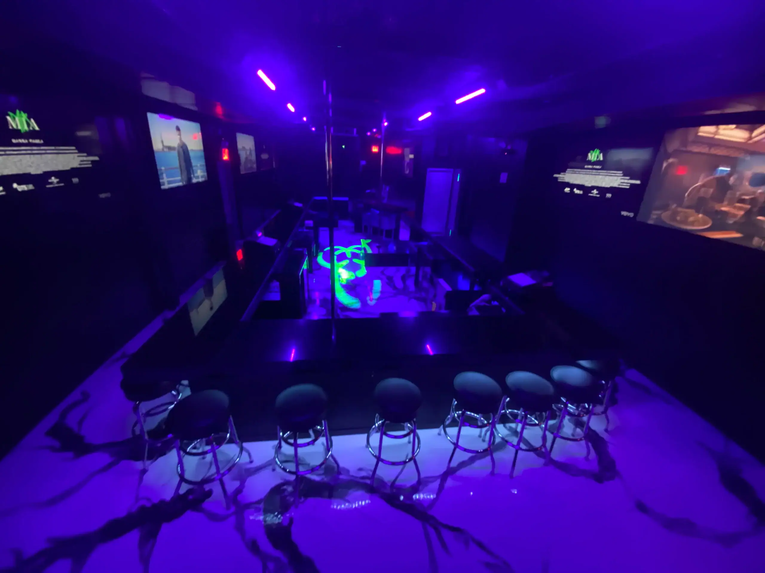 A dimly lit strip club with purple neon lights, a row of barstools facing a mirrored stage with a dance pole, and multiple TV screens on the dark walls displaying various images.