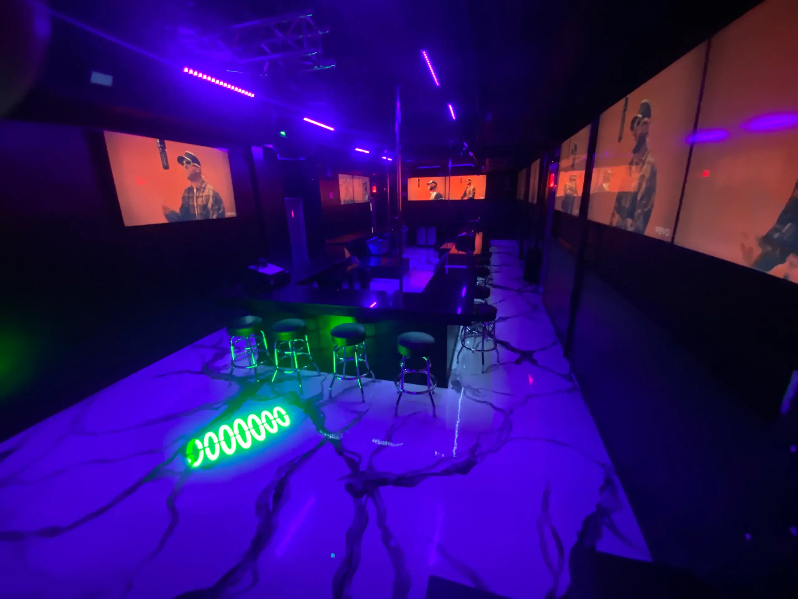 A dimly lit strip club with purple neon lighting, bar stools around a square bar, and large screens on the walls displaying people. The marble-look floor reflects the vibrant lights.