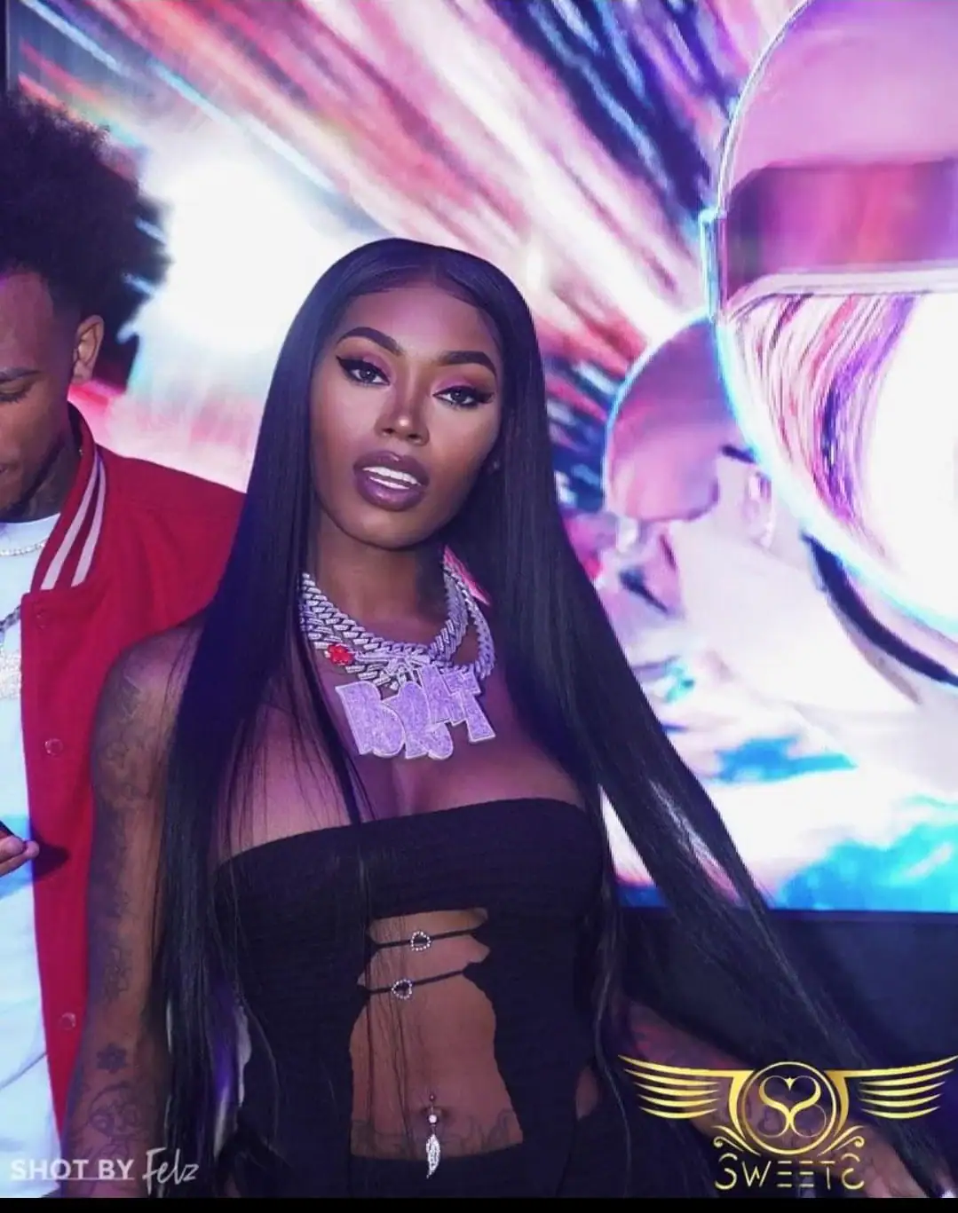 A woman with long straight black hair and bold makeup poses in a black strapless top, layered silver necklaces, and a "DOLL" pendant—channeling Strip Club glamour. A colorful digital background and another person appear behind her; the Sweets logo is in the corner.