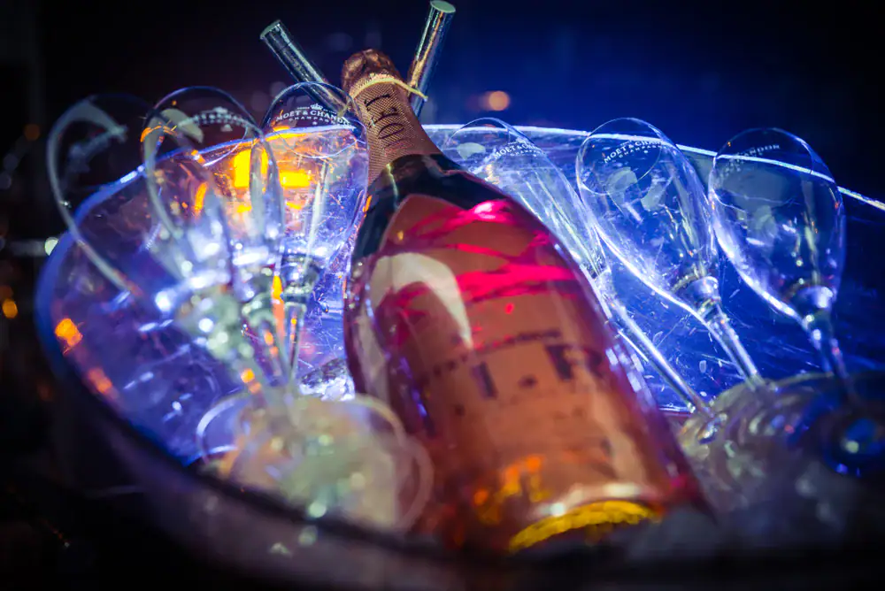 A bottle of champagne lies on ice in a bowl, surrounded by several empty champagne flutes, illuminated by colorful blue and orange lights.