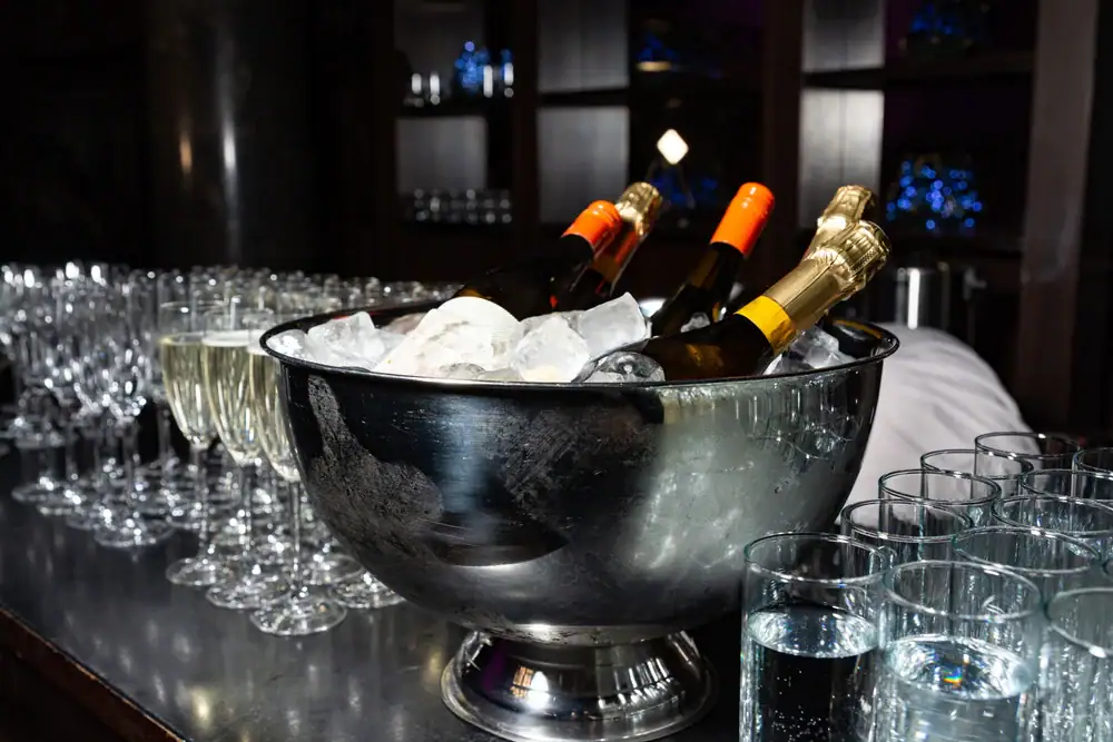 Several bottles of champagne rest in a large metal ice bucket on a table, surrounded by rows of empty champagne flutes and glasses of water, ready for a lively strip club celebration or party.
