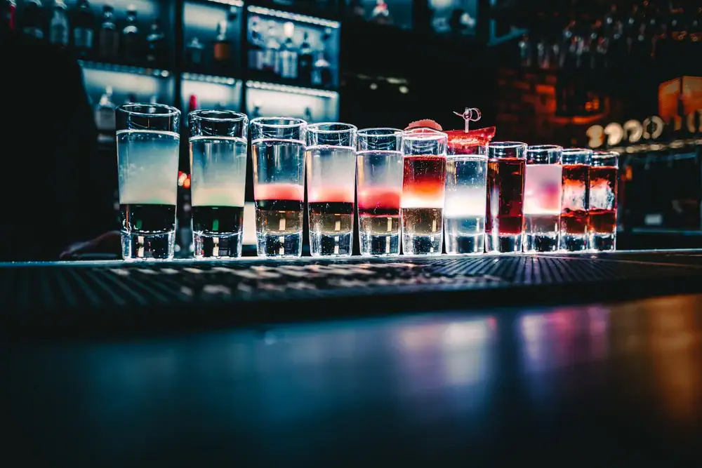 A row of colorful layered cocktails in shot glasses sits on a bar counter, illuminated by blue and white lighting, with bottles and glasses visible on shelves in the background.